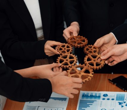 Closeup top view businesspeople hand holding gear and join together over meeting table with financial report papers. Cohesive group of office workers holding cog wheel as synergy harmony concept.